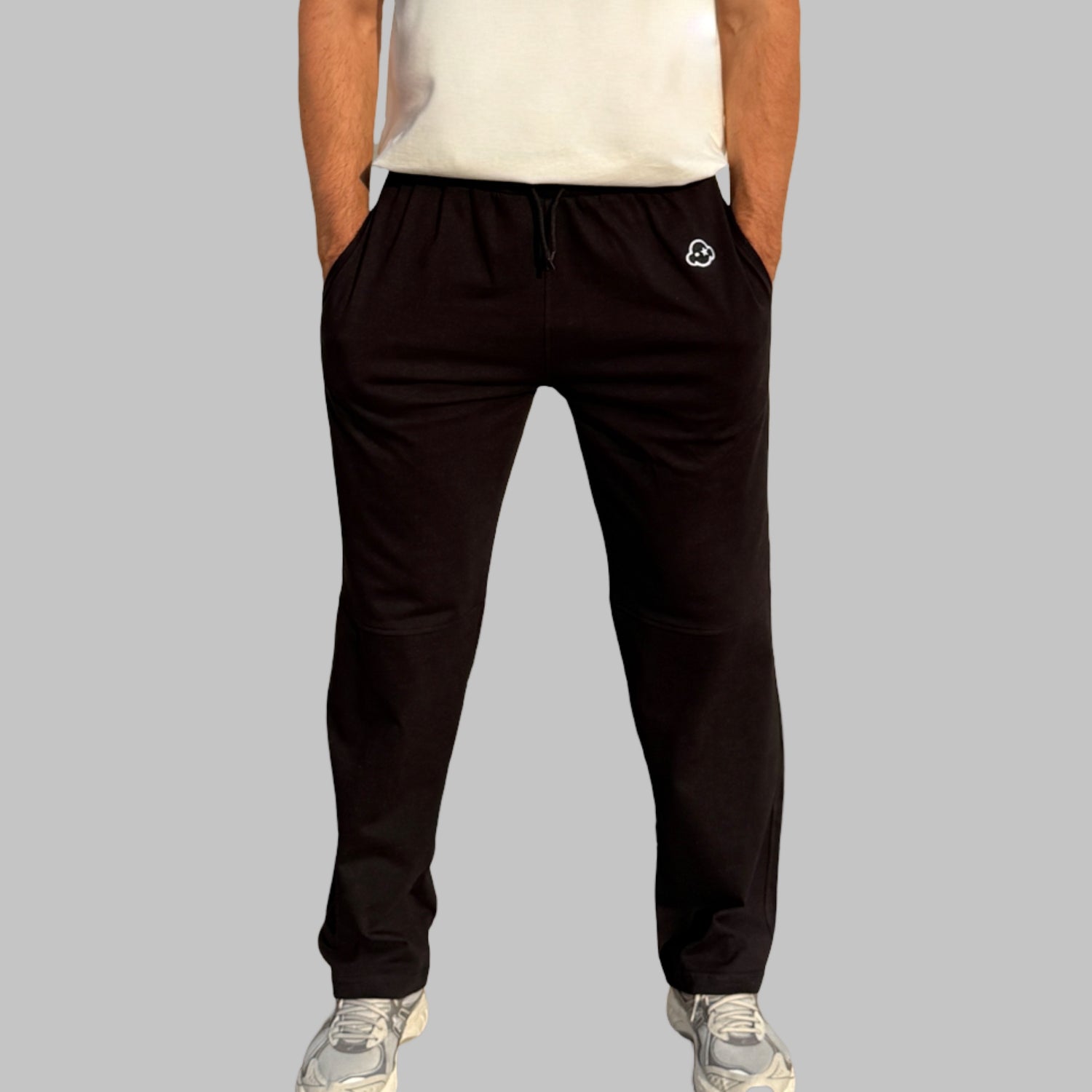 A person standing wearing black activewear pants with a logo on the leg and a white t-shirt. White cotton activewear pants with a small black logo on the left leg, featuring a relaxed fit and drawstring waist. 100% Cotton available in Dubai same day delivery best of summer and winter casual streetwear unisex men and women shorts and t-shirts available in UAE fast delivery and same day delivery available Pants joggers jogger pants