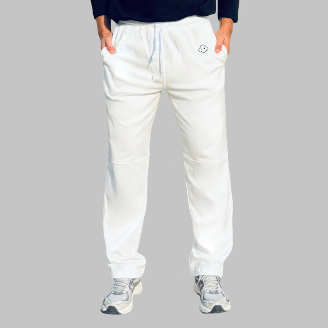 A person standing, wearing white activewear pants with a logo on the left leg and a pair of sneakers. White cotton activewear pants with a small black logo on the left leg, featuring a relaxed fit and drawstring waist. 100% Cotton available in Dubai same day delivery best of summer and winter casual streetwear unisex men and women shorts and t-shirts available in UAE fast delivery and same day delivery available Pants joggers jogger pants