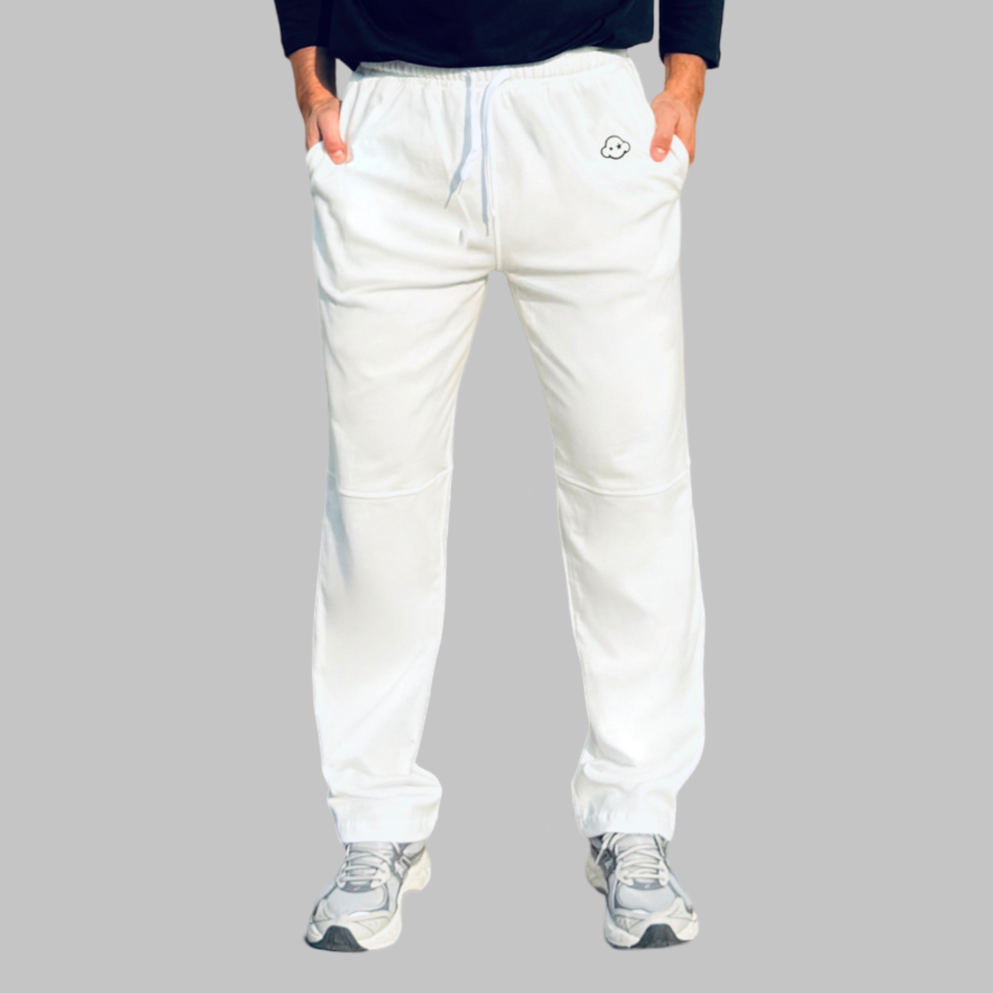 A person standing, wearing white activewear pants with a logo on the left leg and a pair of sneakers. White cotton activewear pants with a small black logo on the left leg, featuring a relaxed fit and drawstring waist. 100% Cotton available in Dubai same day delivery best of summer and winter casual streetwear unisex men and women shorts and t-shirts available in UAE fast delivery and same day delivery available Pants joggers jogger pants